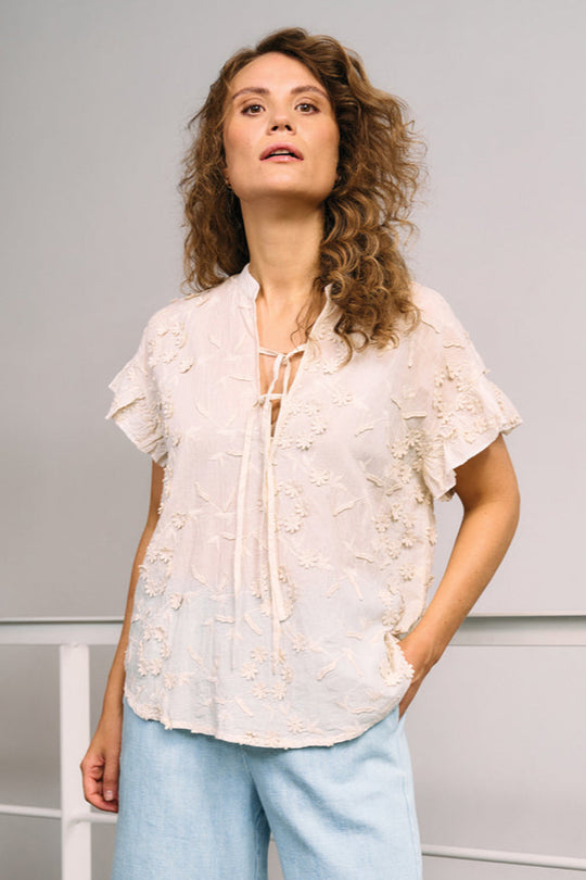 La Rouge Britt Blouse i let bomuld med 3D-blomster, flæseærmer og V-udskæring. Feminin og luftig sommerbluse i creme.