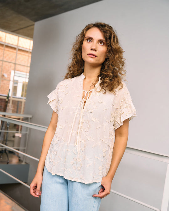 La Rouge Britt Blouse i let bomuld med 3D-blomster, flæseærmer og V-udskæring. Feminin og luftig sommerbluse i creme.